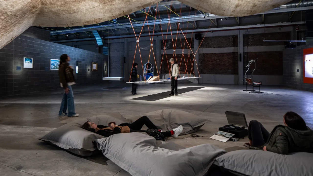 Ampio spazio espositivo moderno con persone che interagiscono e si rilassano: alcuni visitatori camminano e osservano opere d’arte alle pareti, mentre altri riposano su grandi cuscini grigi in primo piano; al centro si distingue un’installazione sospesa con corde arancioni e una piattaforma.