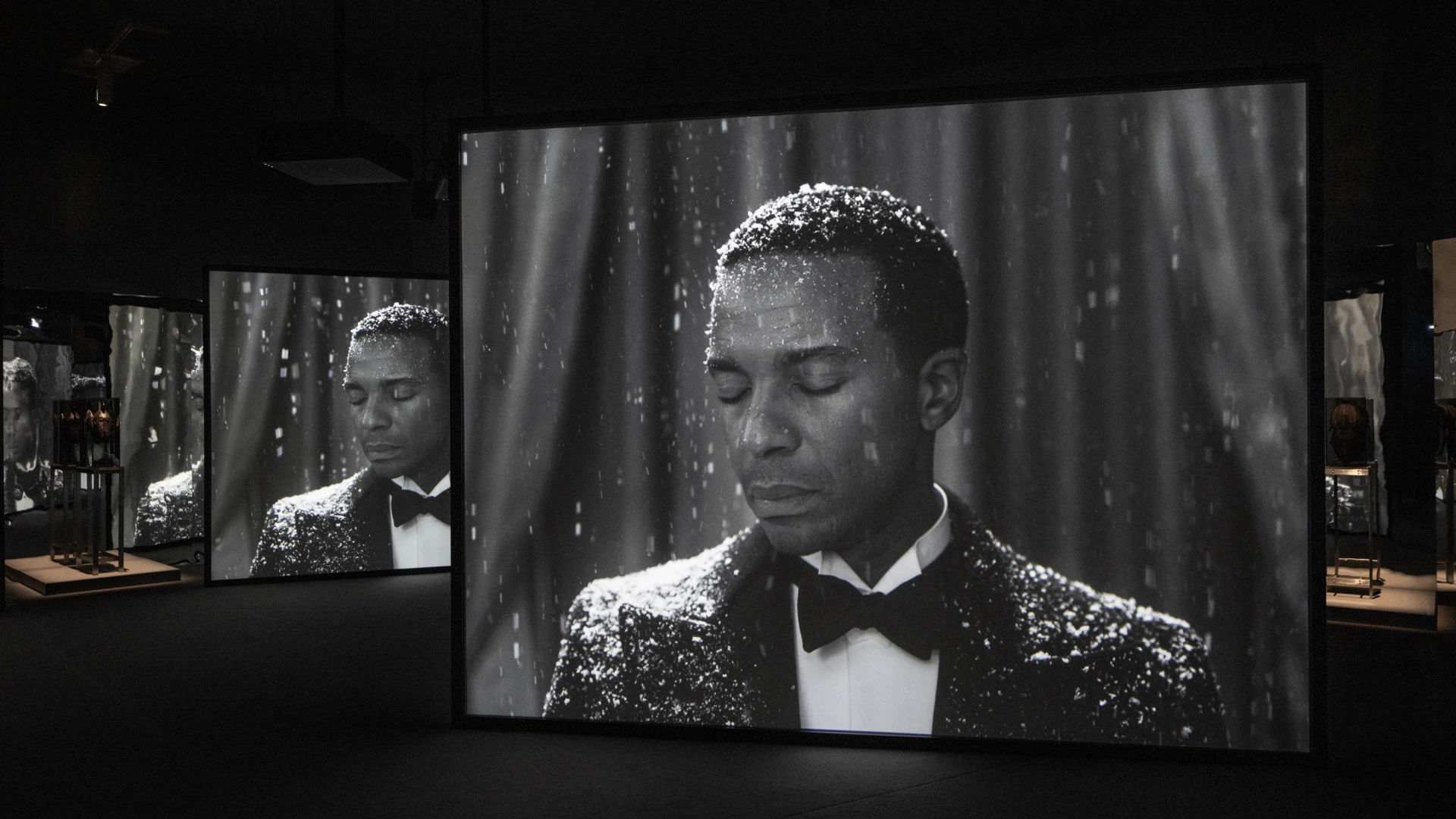 L'immagine raffigura l'installazione "Once Again… (Statues Never Die)" di Isaac Julien alla Tate Britain: schermi multipli mostrano un uomo afroamericano in smoking nero, con gocce d'acqua sul viso, in un ambiente buio da galleria d'arte. © Henrik Kam 2023.