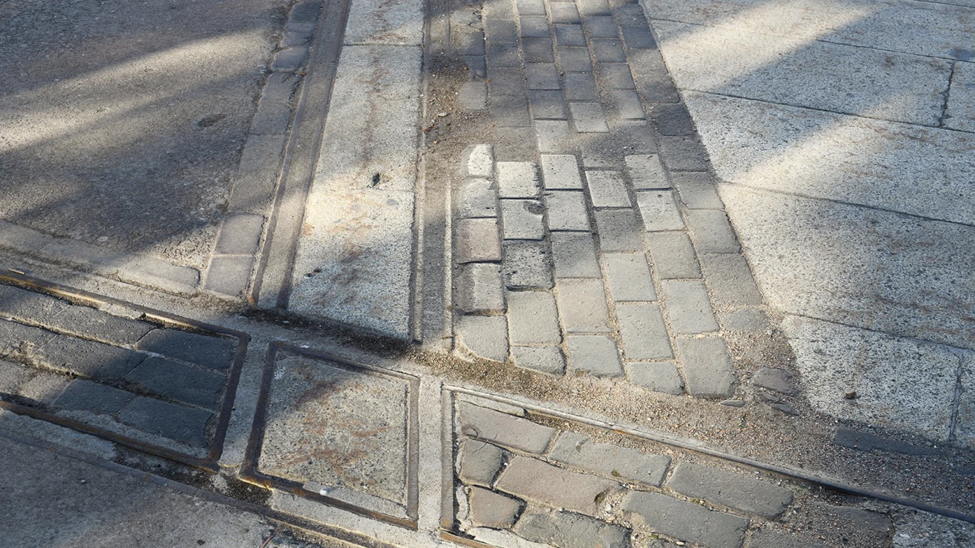 Dettaglio di un marciapiede urbano con binari tramviari dismessi, lastre di cemento e mattoni consumati, attraversati da ombre diagonali che creano geometrie di luce e ombra sulla superficie.