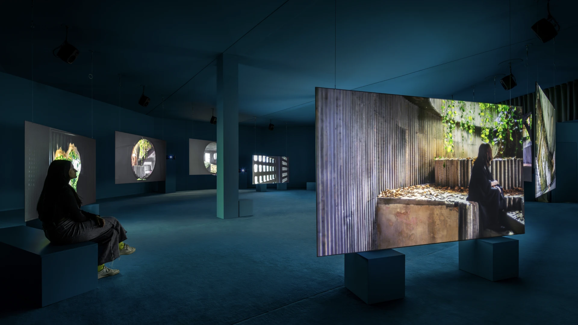 una visitatrice sedua su un cubo osserva le proiezioni di una delle sale delle mostra Museum Dreams di Isaac Julien. Nove schermi posizionati a cerchio proiettano l'opera "Lina Bo Bardi – A Marvellous Entanglement". Le luci dei proiettori illuminano la sala creando un'atmosfera suggestiva e teatrale
